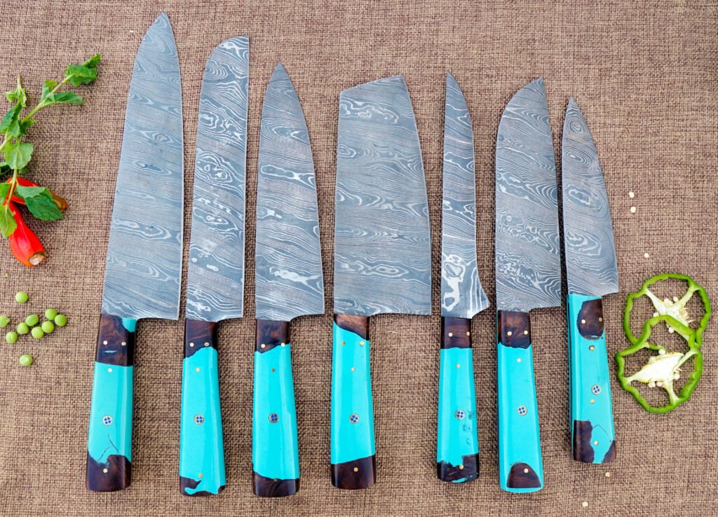 A 7-piece handmade Damascus steel kitchen knife set displayed on a textured background. The blades feature intricate wavy patterns and are paired with vibrant turquoise resin and dark wood hybrid handles. The set includes various shapes like a Cleaver, Chef’s Knife, and Paring Knife, shown alongside fresh green peppers and herbs.